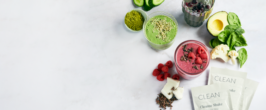 A styled flat-lay of healthy smoothies and wellness ingredients on a white marble surface. Visible items include a green smoothie, a pink smoothie topped with berries, sliced avocado, spinach, cauliflower, cucumber, matcha powder, and several packets labeled CLEAN. A styled flat-lay of healthy smoothies and wellness ingredients on a white marble surface. Visible items include a green smoothie, a pink smoothie topped with berries, sliced avocado, spinach, cauliflower, cucumber, matcha powder, and several packets labeled CLEAN.