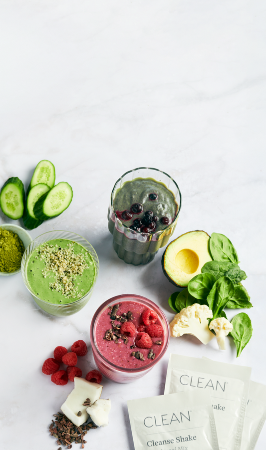 Two prepared smoothies and several supplement packets labeled CLEAN, arranged with fresh ingredients including cucumber slices, green powder, berries, avocado, spinach, cauliflower, and raspberries on a light marble surface. Two prepared smoothies and several supplement packets labeled CLEAN, arranged with fresh ingredients including cucumber slices, green powder, berries, avocado, spinach, cauliflower, and raspberries on a light marble surface.