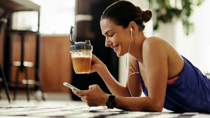 Woman laying on her stomach drinking a protein shake with headphones in and looking at her cell phone