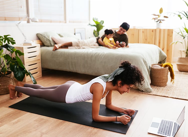woman on a yoga mat in front of a laptop at home with her partner and kid on the bed behind her. 