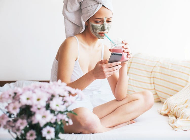 Woman sitting on bed drinking smoothie looking at phone while wearing a detox mask