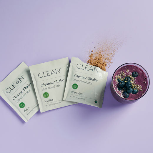Three packets of Cleanse Shake Mix in different flavors: vanilla, chocolate, and plain, with a prepared smoothie topped with granola and blueberries.