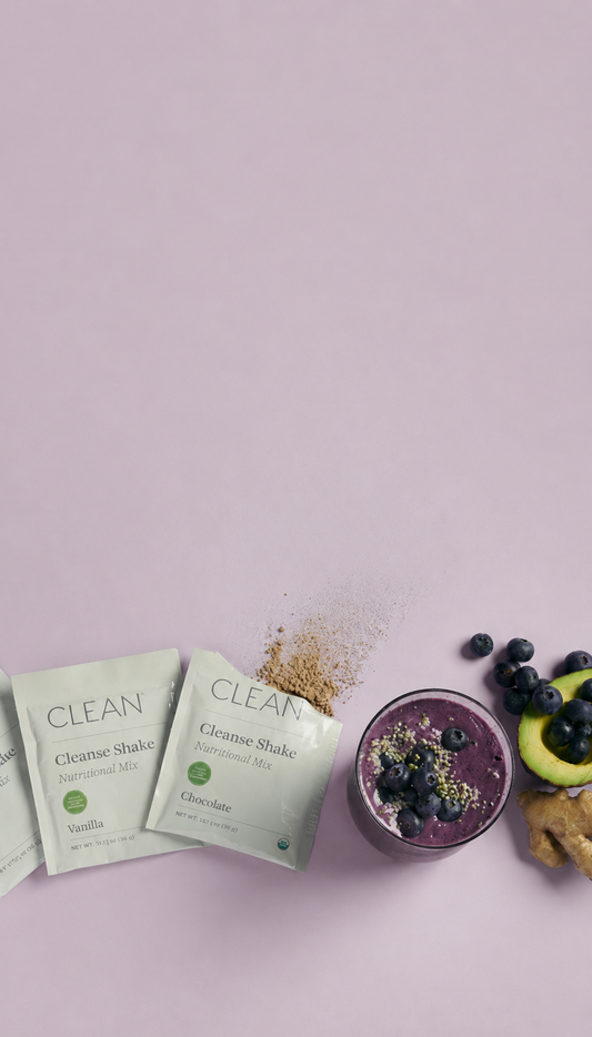Three packets of CLEAN Cleanse Shake powder arranged on a light pink background, next to a purple smoothie bowl topped with blueberries and seeds, with sliced avocado, blueberries, and ginger nearby.
