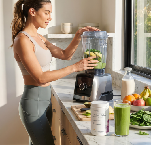 Woman preparing a green smoothie in a blender with ingredients and a supplement container on the counter.