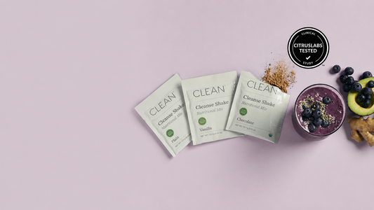Three packets of Citruslabs Clean Clenche Shake powder are arranged on a light pink background next to a prepared purple smoothie with blueberries, a slice of avocado, and ginger. A black circular 'Citruslabs Tested' badge is in the top right corner.