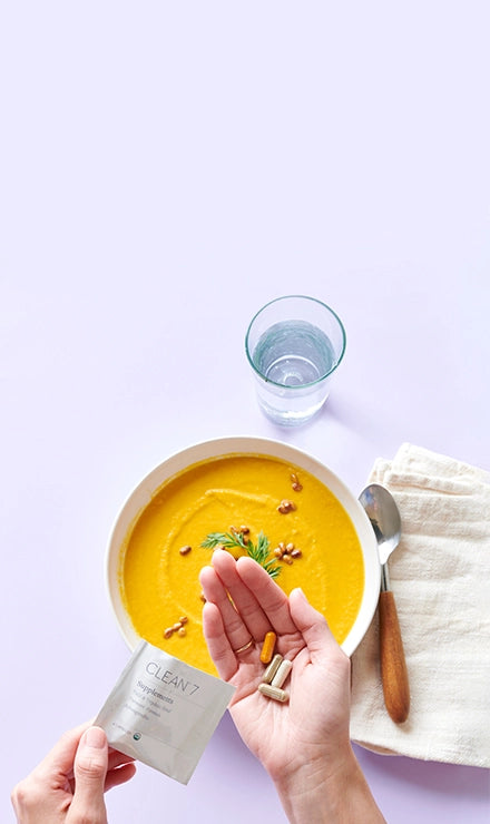 Person holding a Clean 7 supplement package with a bowl of soup and a glass of water on a light purple background